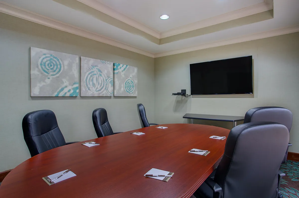 An interior shot of a conference room with a large wooden table, black leather chairs, abstract art on the wall, and a flat-screen TV. The atmosphere is focused and modern, suited for meetings or presentations.