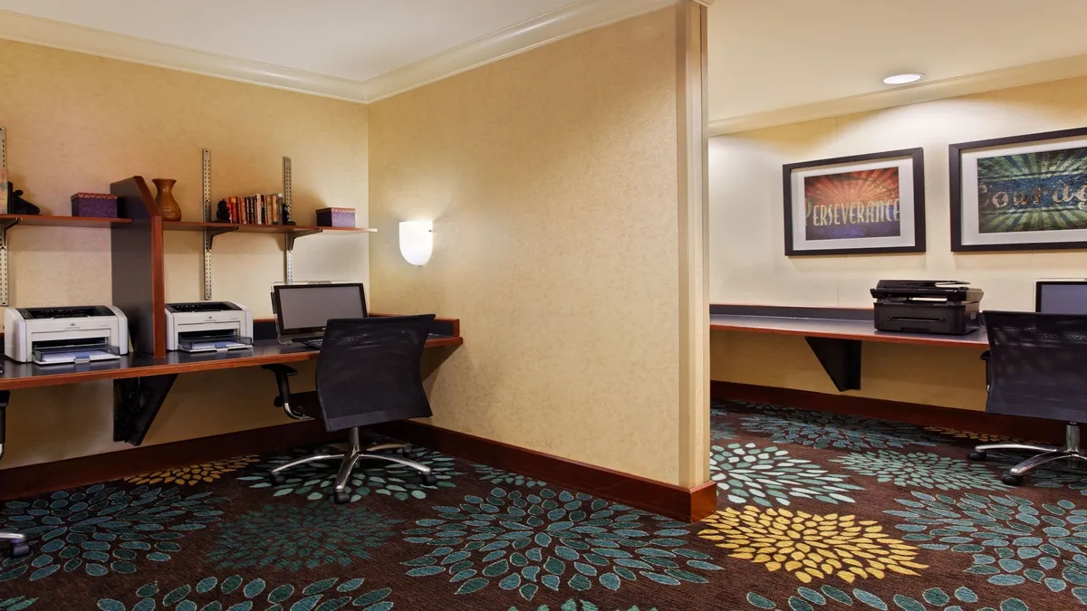 This image shows a modern business center with multiple desks equipped with computers, a colorful patterned carpet, and motivational posters on the wall. The environment is organized and styled to promote productivity, making it ideal for guests looking to work or study during their stay.
