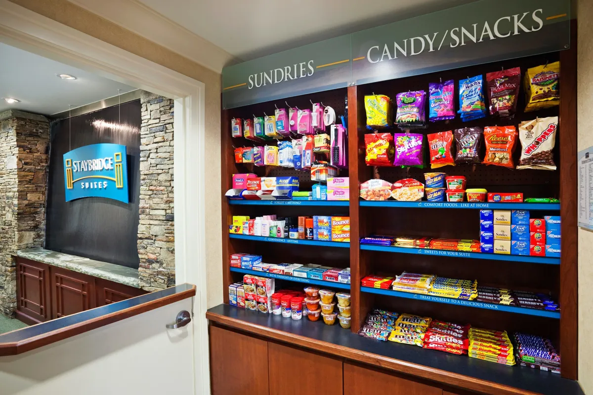 A casual sundries shelf beside a stone wall and reception area, displaying a range of snacks, quick meals, and essentials. The setup invites guests to grab ingredients or treats to enjoy in their own kitchen or dining area.