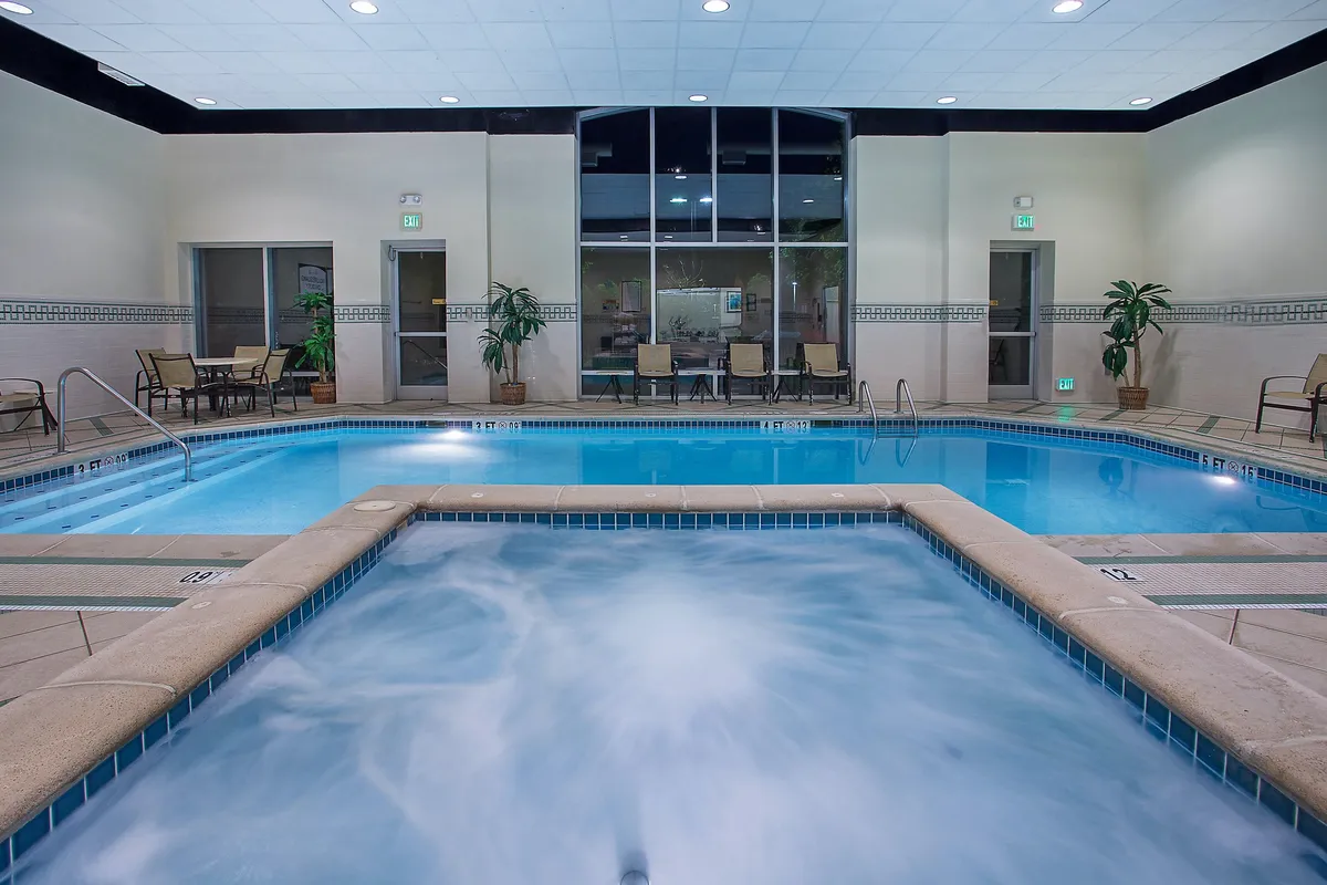 This image features a heated indoor pool and a hot tub, flanked by a surrounding seating area. Large windows let in natural light and provide a serene, inviting atmosphere. Guests are shown relaxing in the hot tub, swimming, and chatting poolside, emphasizing the space's dual focus on relaxation and social interaction.
