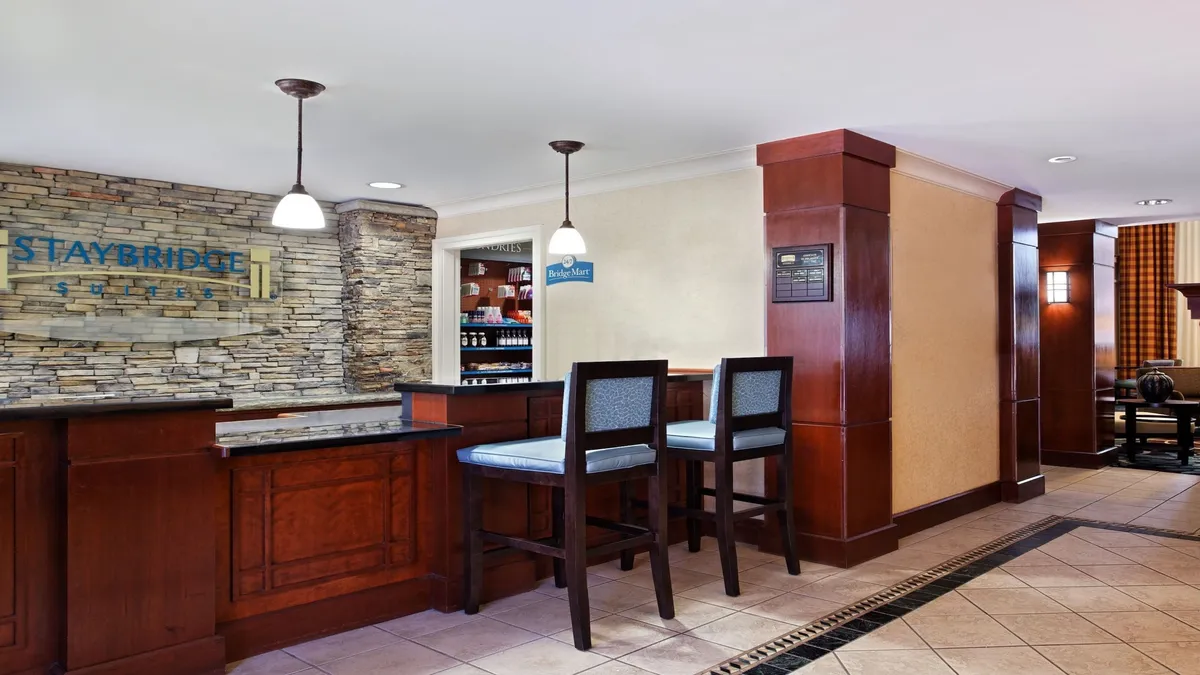 The image captures the lively reception area, featuring a stone wall, a welcoming reception desk, and a bar-style seating area. Several guests are seen checking in, chatting by the bar, and enjoying a casual coffee, reflecting the social, communal atmosphere described in the section.