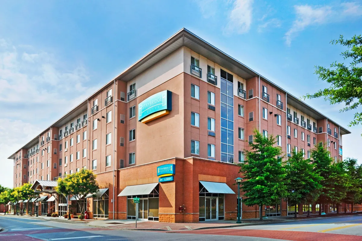 This image shows the hotel’s exterior facade, complete with clear signage and trees lining the street. People are seen strolling by, sitting on benches, and exploring nearby shops, creating a lively and inviting street scene right across from the convention center.