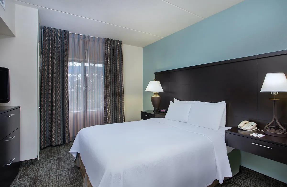A tranquil bedroom featuring a plush queen bed, a nightstand with an elegant lamp, floor-length curtains framing a window, and a wall-mounted television. The color palette is soothing, and subtle decor accents add to the restful ambiance.