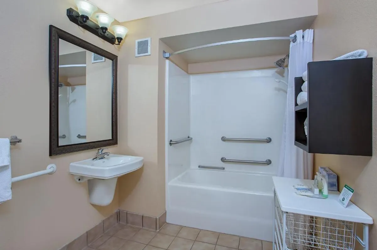 This image showcases a spotless, well-lit bathroom featuring a modern sink, expansive mirror, plush towels neatly arranged on a shelf, and a pristine bathtub—inviting guests to unwind and refresh in comfort.