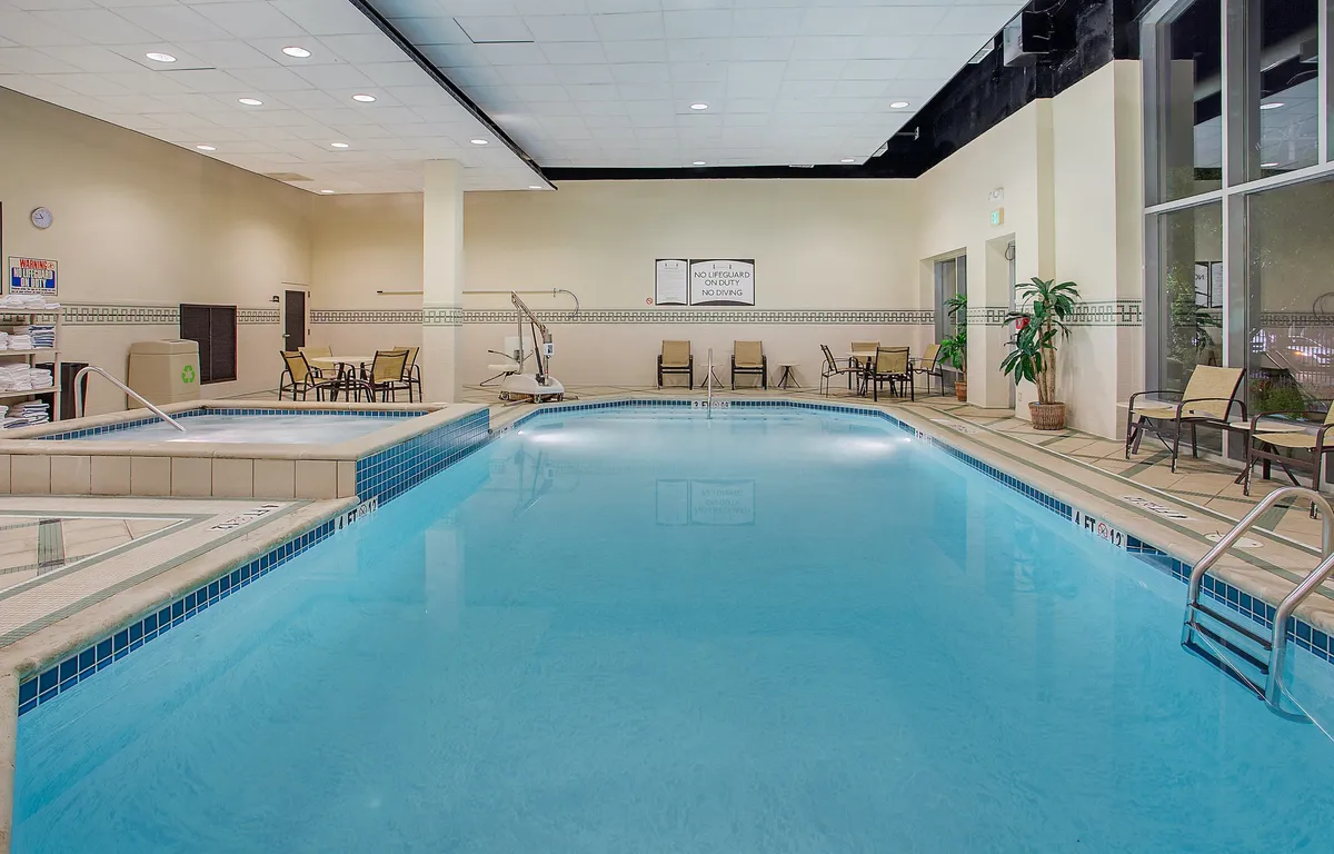 This image features a tranquil indoor swimming pool area with lounge chairs, lush indoor plants, and a wall clock. The pool is spacious and inviting, framed by comfortable seating for guests to relax, read, or socialize. The clean, airy design and abundance of natural light create a refreshing retreat after a day of urban adventures.