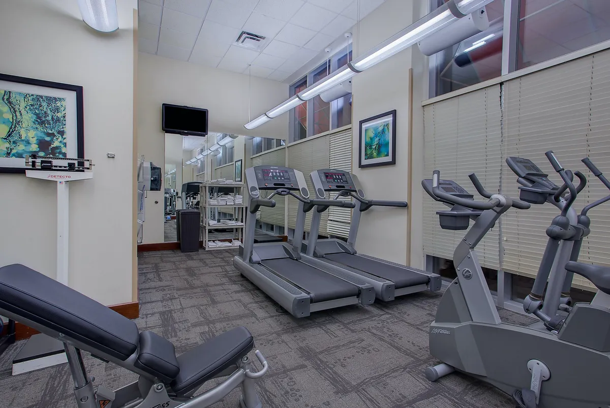 A spacious fitness center featuring a row of treadmills, an elliptical machine, weight bench, large mirrors, and colorful artwork. The room is clean and inviting, encouraging an energetic workout any time of day.