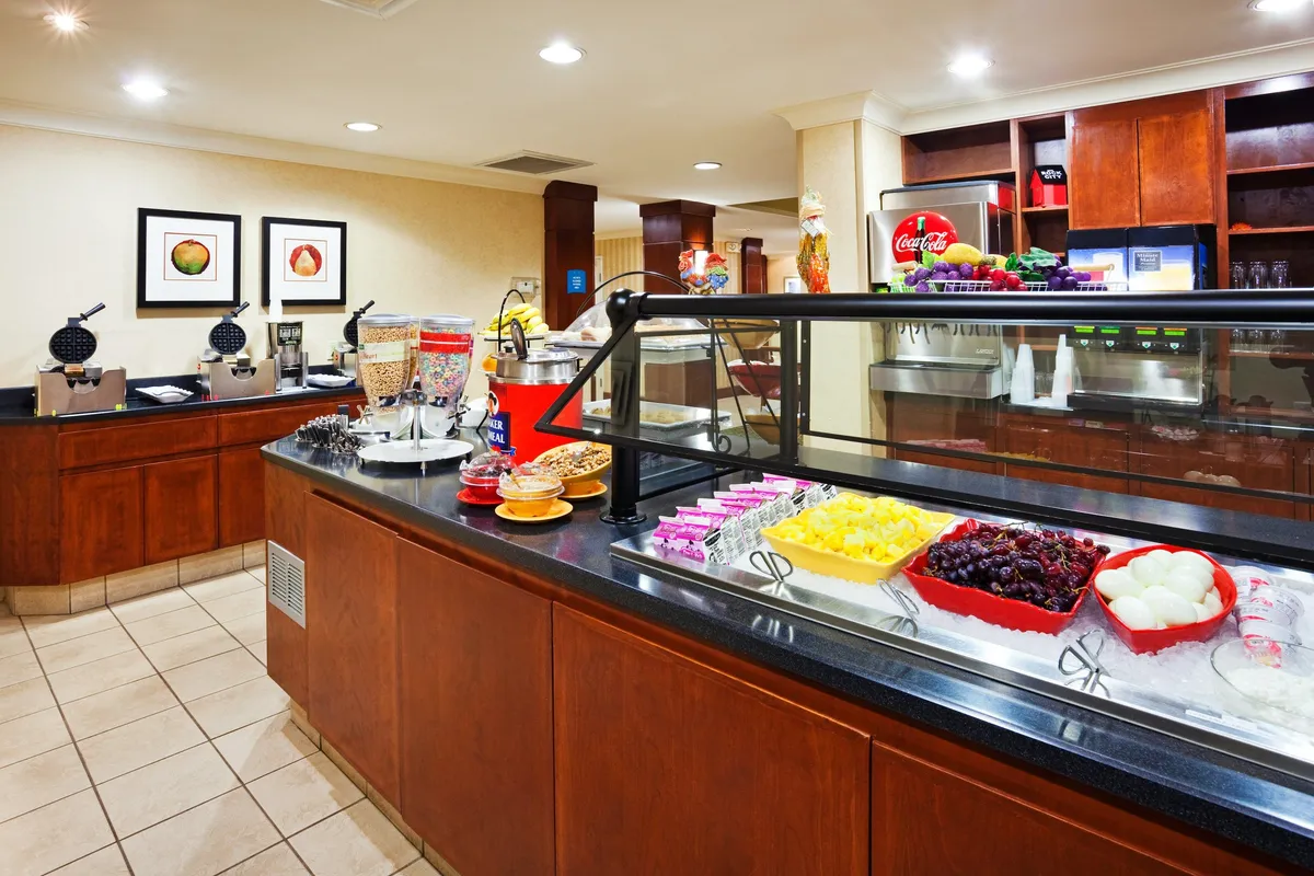 A bright, inviting breakfast bar featuring a spread of fruit, beverages, and dessert options. The self-service layout showcases a counter with stools, encouraging guests to enjoy a quick meal or snack in a communal setting.