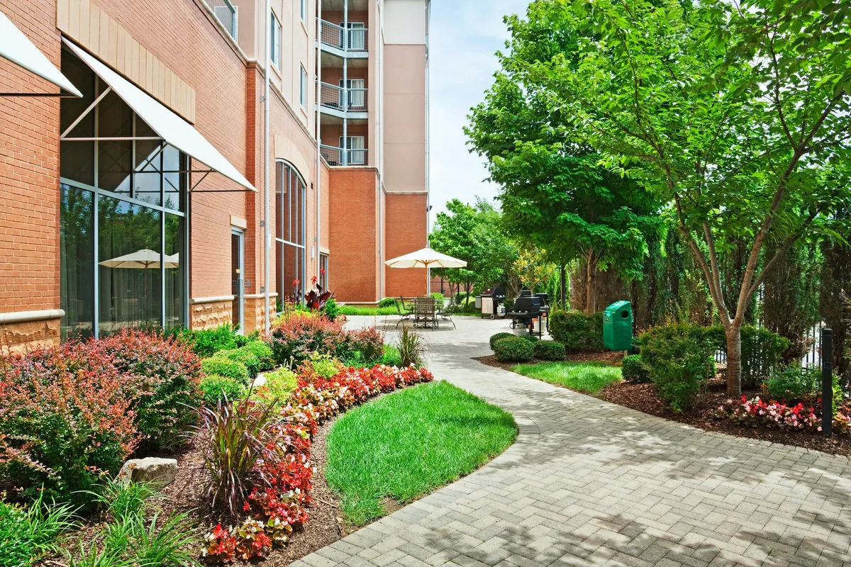 An outdoor seating area surrounded by colorful flower beds and trees creates a peaceful setting for social gatherings. The space is well-designed for group barbecues or quiet moments enjoying nature, with comfortable seating and easy access to the building.